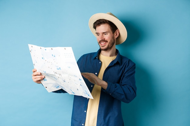 Knappe glimlachende toerist in strohoed die kaart bekijkt, reisweg kiest, een vakantie plant, die zich op blauwe achtergrond bevindt.