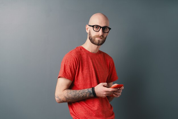 Knappe haarloze man met baard, bril, rood T-shirt, sms'en terwijl hij smartphone vasthoudt