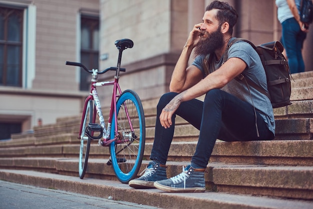 Knappe hipster reiziger met een stijlvolle baard en tatoeage op zijn armen gekleed in vrijetijdskleding, zittend op de trap, met behulp van de telefoon, rustend na een fietstocht.