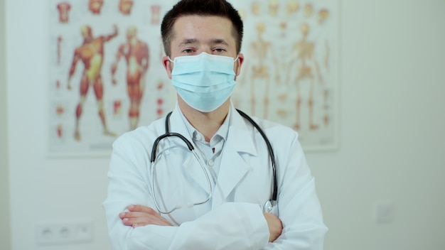 Knappe huisarts in medisch kantoor. Succesvolle arts met beschermend masker en witte laboratoriumjas die naar de camera kijkt en lacht in de ziekenhuiskliniek.