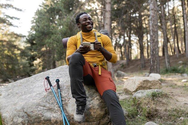 knappe jonge Afrikaanse Amerikaanse man met rugzak op de berg hij heeft een mobiele telefoon en een beker hij zit op de rots concept van trekking reiziger rugzak