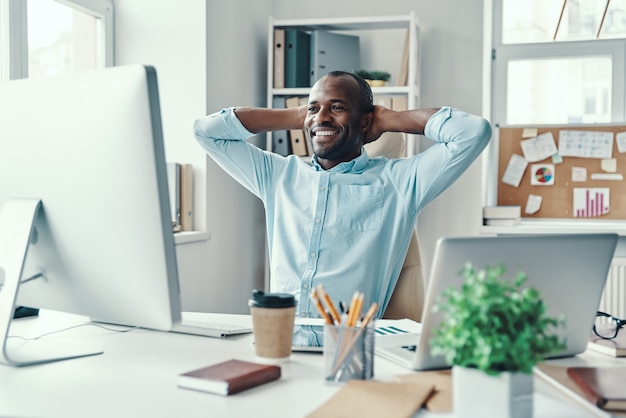 Knappe jonge Afrikaanse man in shirt die handen achter het hoofd houdt en glimlacht terwijl hij op kantoor werkt