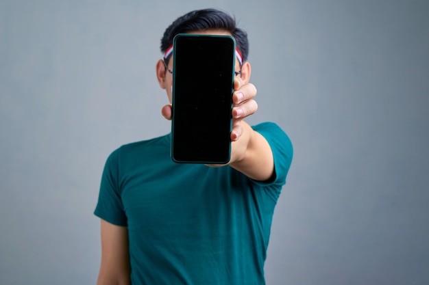 Knappe jonge aziatische man in casual tshirt weergegeven: mobiele telefoon met leeg scherm aanbevelen van mobiele app geïsoleerd op grijze achtergrond indonesische onafhankelijkheidsdag viering concept