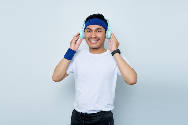 Knappe jonge Aziatische man sportieve fitness trainer instructeur in blauwe hoofdband en witte tshirt maakt plezier luisteren naar muziek met koptelefoon geïsoleerd op witte achtergrond