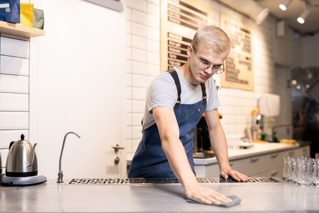 Knappe jonge barista of ober in werkkleding zijn werkplek afvegen met natte stofdoek tijdens de voorbereiding op het werk aan het begin van de dag