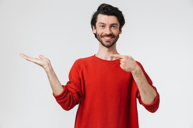 Knappe jonge bebaarde brunette man met trui permanent over wit, wijzende vinger op kopie ruimte