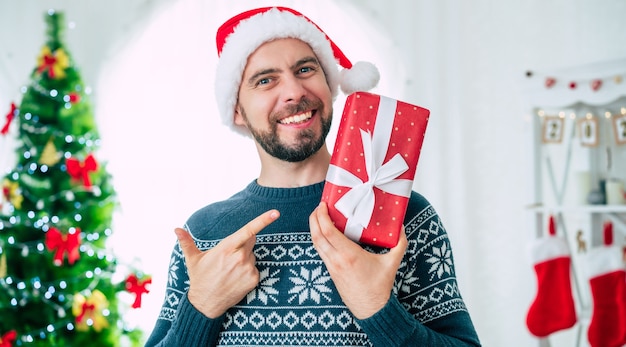 knappe jonge bebaarde lachende man in een kerstmuts met een kerstcadeau