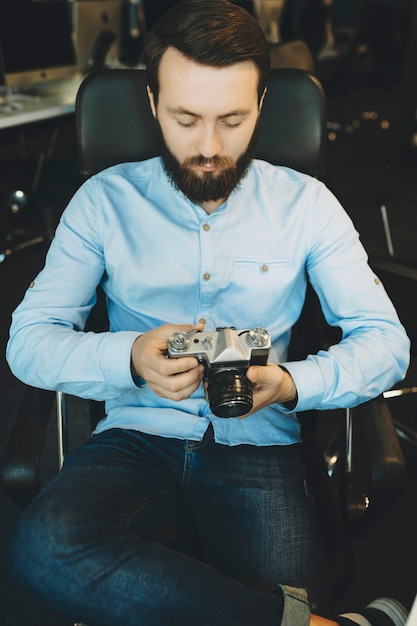 Knappe jonge bebaarde man in blauw shirt en spijkerbroek comfortabel zittend op een stoel met gekruiste benen en aandachtig kijken naar camera in handen met werkplek op onscherpe achtergrond