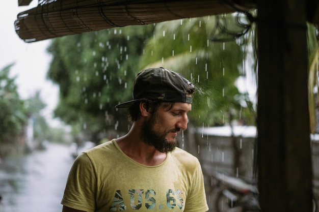 Knappe jonge, bebaarde man in een pet onder de tropische regen.
