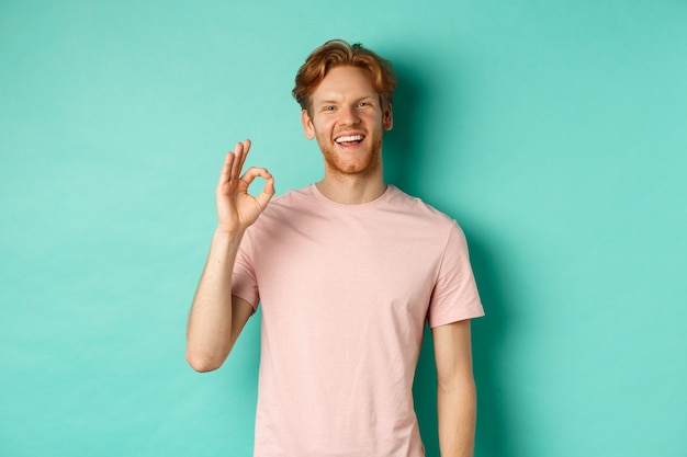 Knappe jonge bebaarde man in t-shirt ok teken tonen, glimlachend met witte tanden en ja zeggen, ben het met je eens, staande op turkooizen achtergrond.