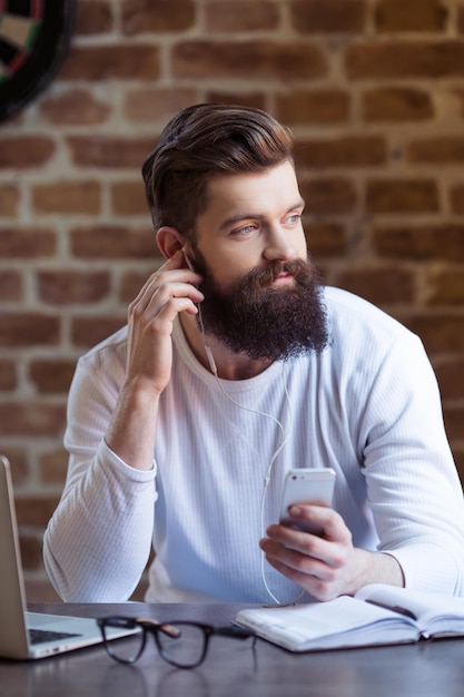 Knappe jonge, bebaarde man in wit sweatshirt luistert naar muziek met behulp van een smartphone en glimlacht na het werken met een laptop