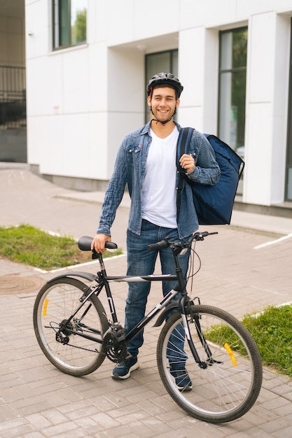 Knappe jonge bezorger in beschermende helm die zich voordeed in de buurt van de fiets in de straat van de stad, kijkend naar de camera. Koerier man met thermo rugzak bezorging van voedsel aan klant