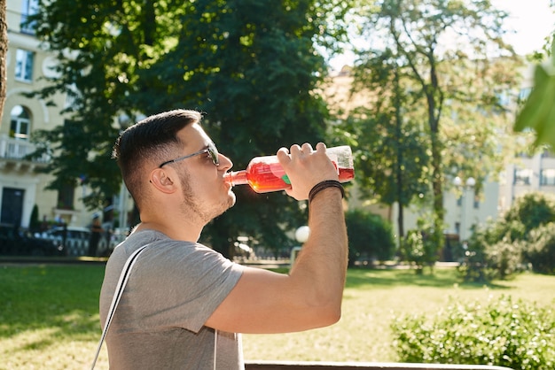 Knappe jonge blanke man met donker haar in zonnebril, grijs t-shirt die een rode koolzuurhoudende drank drinkt uit een fles