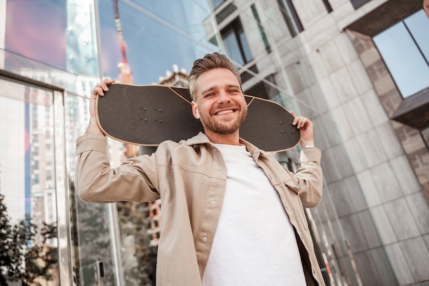 Knappe jonge blonde glimlachende man met skateboard op schouders op straat in de stad tussen glazen gebouwen. Draagt een denim-outfit. Sportieve stijlvolle skateboarder die buiten aan extreme truc denkt.