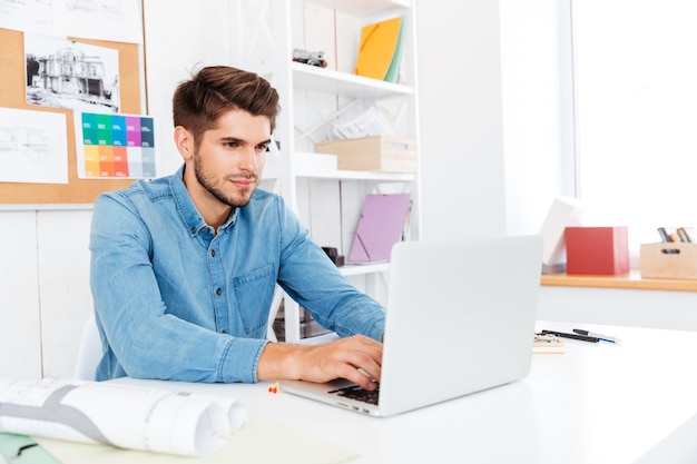 Knappe jonge casual zakenman die met laptop werkt en aan tafel zit op kantoor