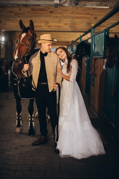 Knappe jonge cowboyman en zijn charmante boho-bruid poseren in een stal met paarden