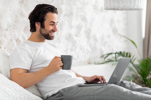 Knappe jonge kerel die laptop in bed gebruikt die koffie drinkt