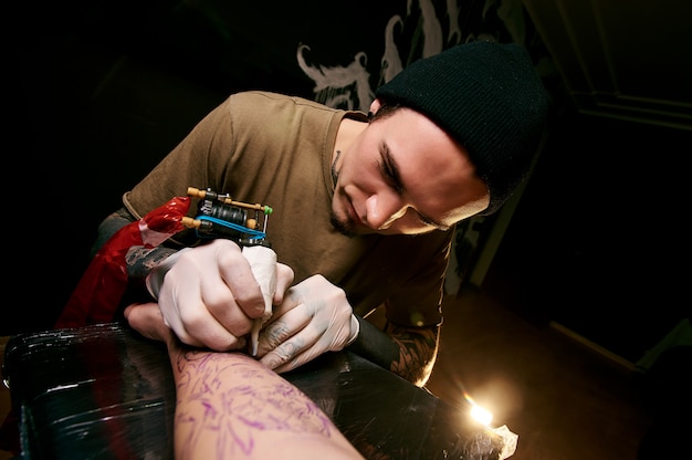 Foto knappe jonge kerel in een zwarte hoed en met tatoeages, slaat een tatoeage op zijn arm, tattoo salon