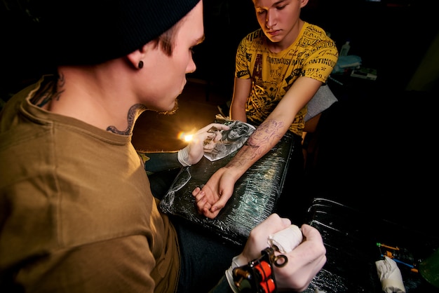 Knappe jonge kerel in een zwarte hoed en met tatoeages, verslaat een tatoeage op zijn arm, tattoo salon