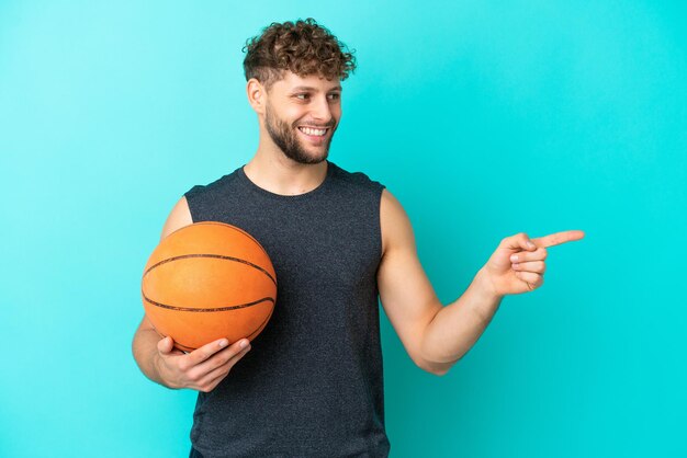 Knappe jonge man die basketbal speelt geïsoleerd op een blauwe achtergrond die met de vinger naar de zijkant wijst en een product presenteert