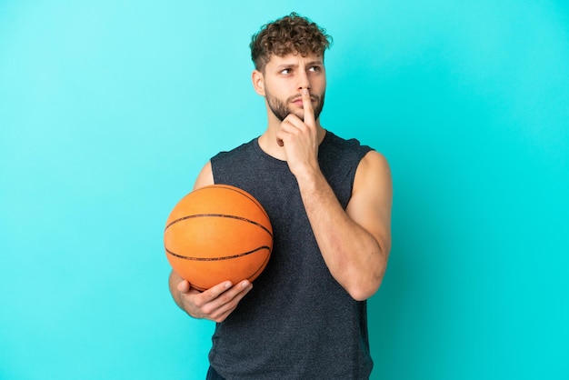 Knappe jonge man die basketbal speelt geïsoleerd op een blauwe achtergrond die twijfelt terwijl hij omhoog kijkt