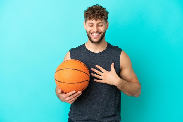 Knappe jonge man die basketbal speelt geïsoleerd op een blauwe achtergrond die veel lacht
