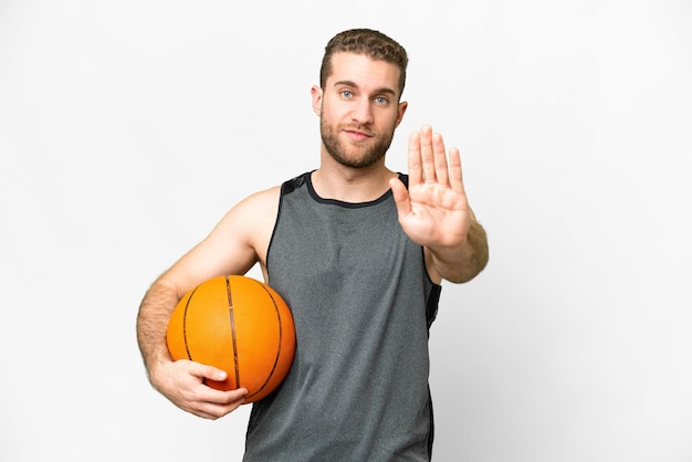 Knappe jonge man die basketbal speelt over een geïsoleerde witte achtergrond die een stopgebaar maakt