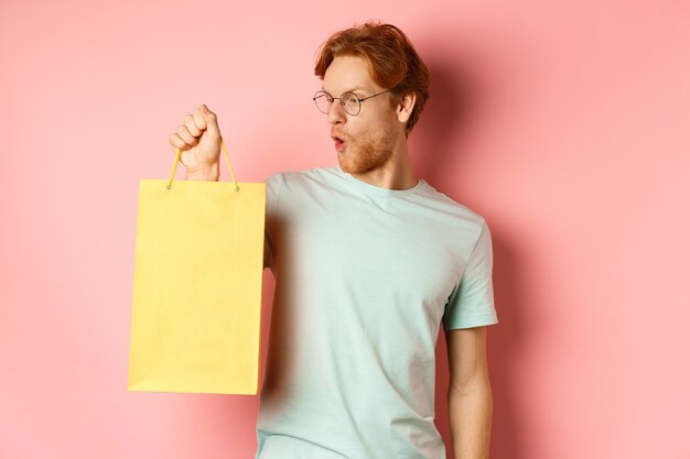 Knappe jonge man die cadeautjes koopt, een boodschappentas vasthoudt en er geamuseerd uitziet, staande over een roze achtergrond