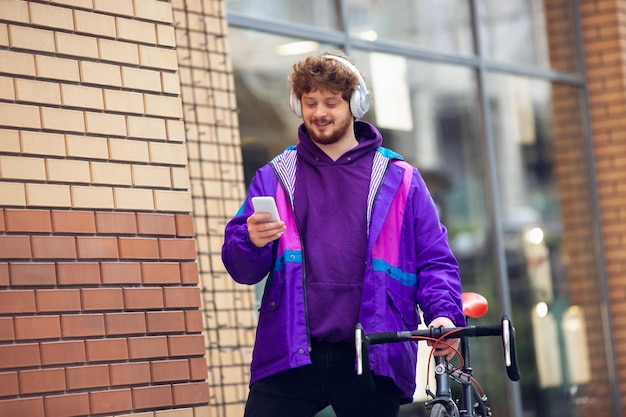 Knappe jonge man die mobiele telefoon en koptelefoon gebruikt terwijl hij naast zijn fiets staat