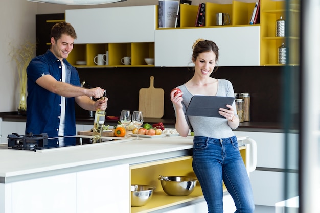 Knappe jonge man die wijnfles opent terwijl zijn vrouw digitale tablet in de keuken gebruikt.