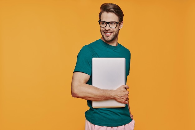 Knappe jonge man in casual kleding die laptop draagt en glimlacht terwijl hij tegen de gele muur staat