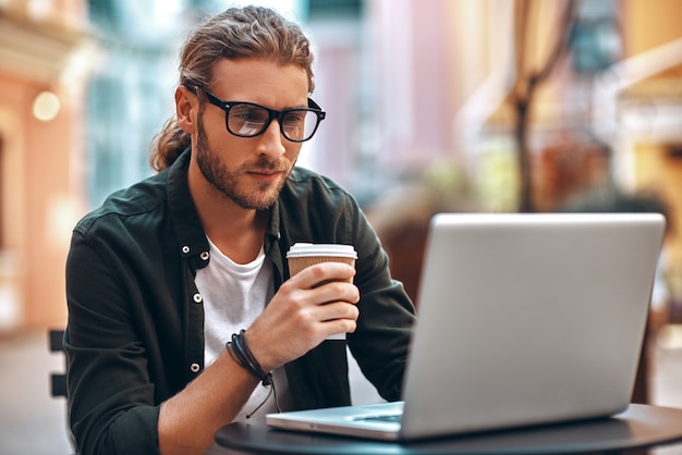 Knappe jonge man in casual kleding die met behulp van de computer werkt en koffie drinkt terwijl hij in café zit