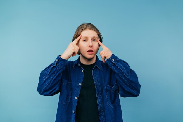 Knappe jonge man in een shirt staat op een blauwe achtergrond toont zijn wijsvingers op zijn slaap