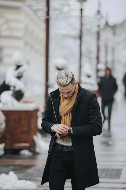 Knappe jonge man in een zwarte winteroutfit met een bruine sjaal die zijn horloge controleert terwijl hij loopt