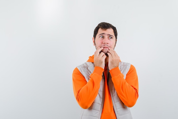 Knappe jonge man in oranje hoodie en wit vest stak vingers over de mond en vol angst geïsoleerd op een witte achtergrond