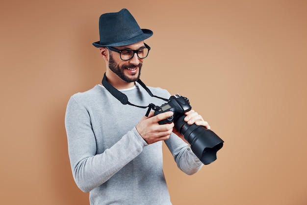 Knappe jonge man in vrijetijdskleding die lacht en de fotocamera aanpast terwijl hij tegen een bruine muur staat