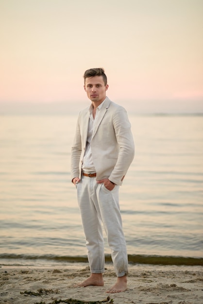 Knappe jonge man in wit pak aan de kust bij zonsondergang
