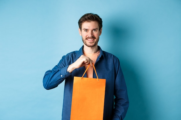Knappe jonge man met baard, glimlachend en boodschappentas tonen, iets kopen in de winkel, staande op blauwe achtergrond.