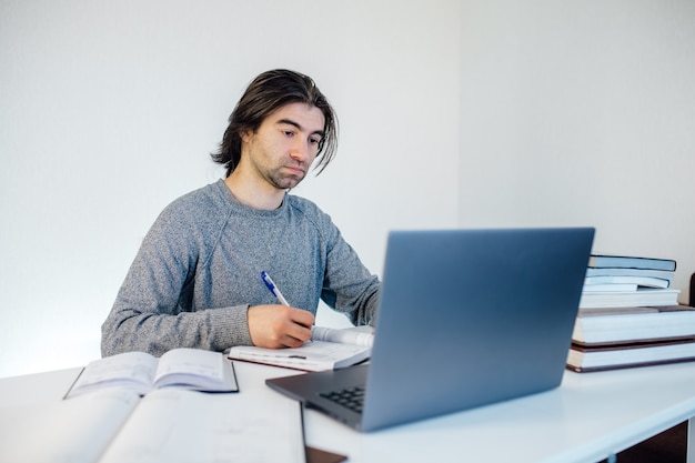 Knappe jonge man met behulp van laptopcomputer thuis. leerling leert aan tafel. Online winkelen, thuiswerk, freelance, online leren, studeren concept. Afstandsonderwijs