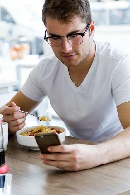 Knappe jonge man met behulp van zijn mobiele telefoon terwijl u geniet van het ontbijt in de keuken.