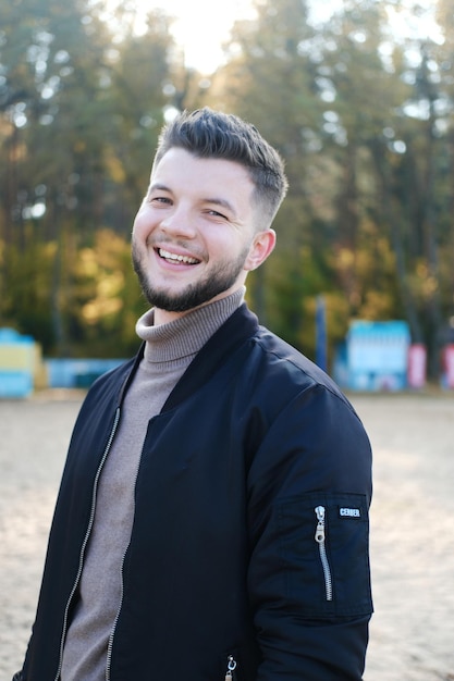 Knappe jonge man met een baard en in een zwarte jas poseren op het strand Man glimlach en kijk naar de camera