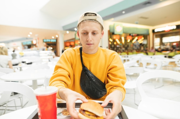 Knappe jonge man met een pet en gele kleren zitten bij de receptie en fast food eten