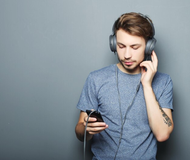 Knappe jonge man met koptelefoon en mobiele telefoon vast te houden terwijl hij tegen een grijze muur staat