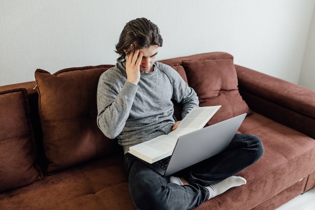 Knappe jonge man met laptopcomputer thuis. Student man leest het boek. Online winkelen, thuiswerk, freelance, online leren, studeren concept. Afstandsonderwijs