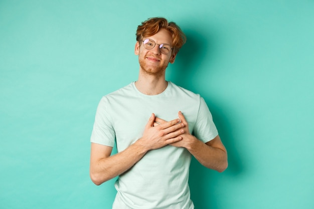 Knappe jonge man met rood haar en bril, hand in hand op het hart en glimlachen, bedankt zeggen, dankbaar en ontroerd voelen, staande over turkooizen achtergrond