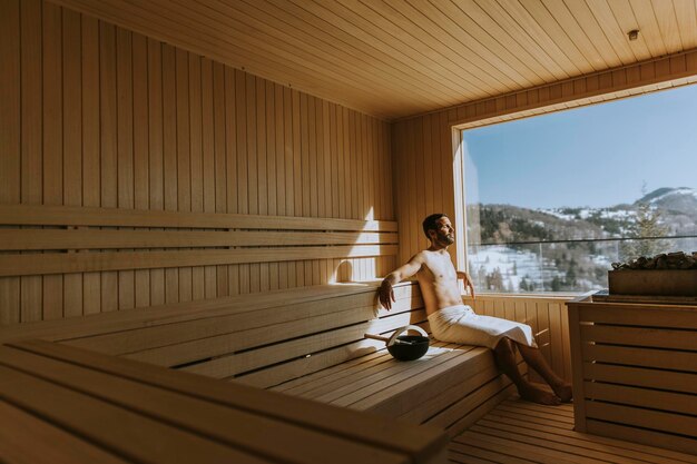 Knappe jonge man ontspannen in de sauna en kijken naar het winterbos door het raam