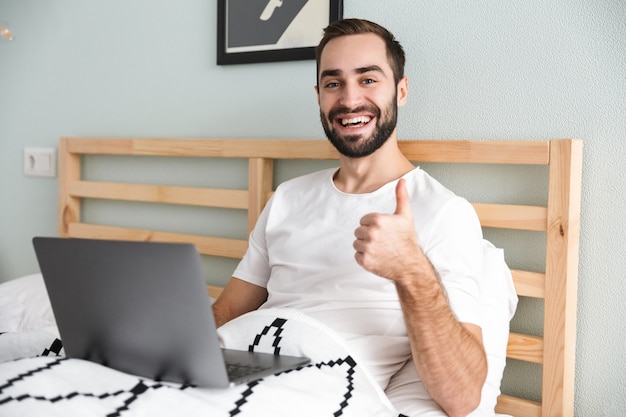 Knappe jonge man tot in bed, werken op laptopcomputer, duimen omhoog