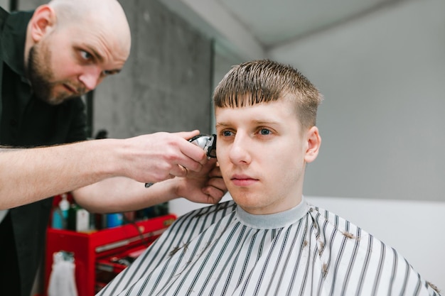 Knappe jonge man zit in een arberwinkelstoel met een serieus gezicht en knipt haar bij zijn kapper