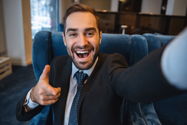 Knappe jonge opgewonden zakenman draagt pak zit in de lobby van het hotel, met behulp van mobiele telefoon, een selfie nemen, wijzen