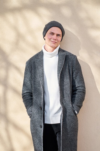 Foto knappe jonge positieve vrolijke hipster stijlvolle man met moderne grijze jas, witte trendy trui en zwarte spijkerbroek die zich in de buurt van beige muur bevindt